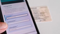 Auskunftsservice_Ausweise Auf dem Bildschirm eines Smartphones sieht man die Eingabemaske, um den Bearbeitungsstand eines Ausweisdokuments abzurufen. Im Hintergrund liegt ein Personalausweis.