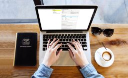 Notebook auf Tisch Zwei Hände tippen an einem Notebook, dass eine Seite auf Service BW geöffnet hat. Links neben dem Notebook liegt ein Notizbuch, rechts neben dem Notebook liegen eine Sonnenbrille und eine Tasse Kaffee.
