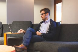 Person am Notebook Ein junger Mann sitzt auf einem Sofa und bedient mit einer Hand ein Notebook das vor ihm steht.
