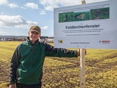 Mann steht auf einem Acker neben einem Informationsschild für Feldlerchenfenster